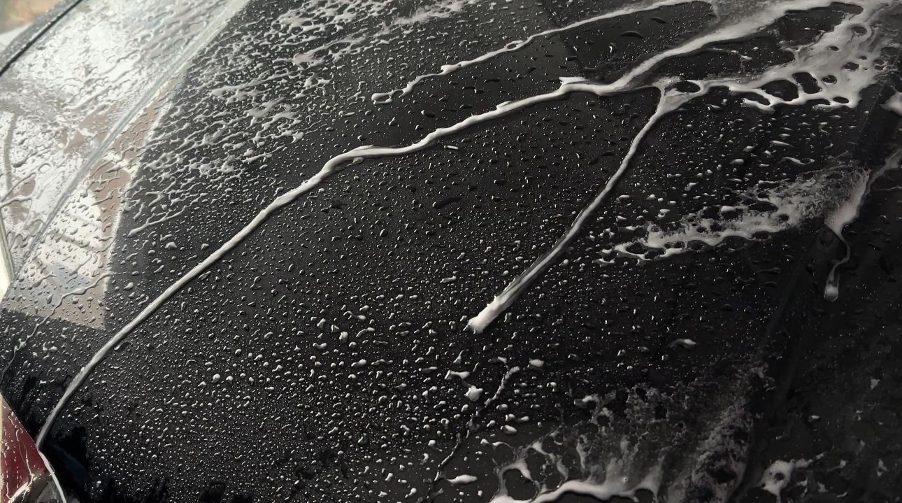 Close-up of a car hood with water droplets and soap streaks during washing.
